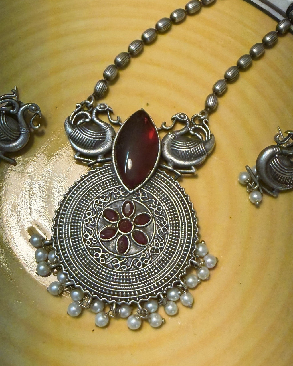 Close-up of circular pendant with red stones, floral design and pearl detailing in oxidised finish