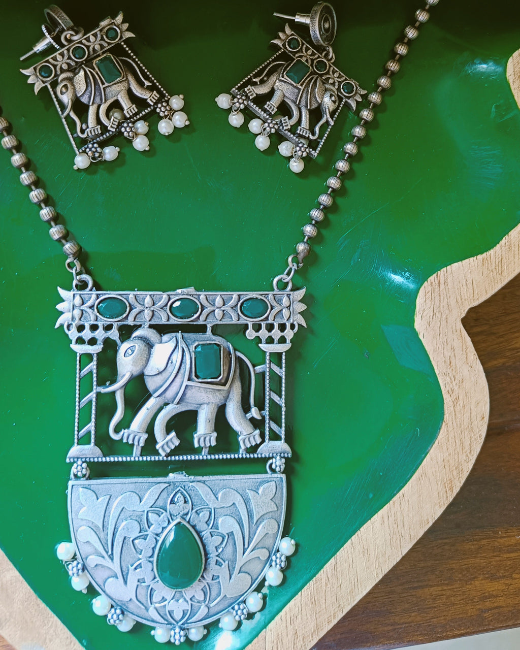 Gajraj Emerald antique oxidised silver elephant pendant necklace and stud earrings set with emerald-green stones displayed on a red background