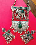 Gajraj Emerald antique oxidised silver elephant pendant necklace and stud earrings set with emerald-green stones displayed on a red background