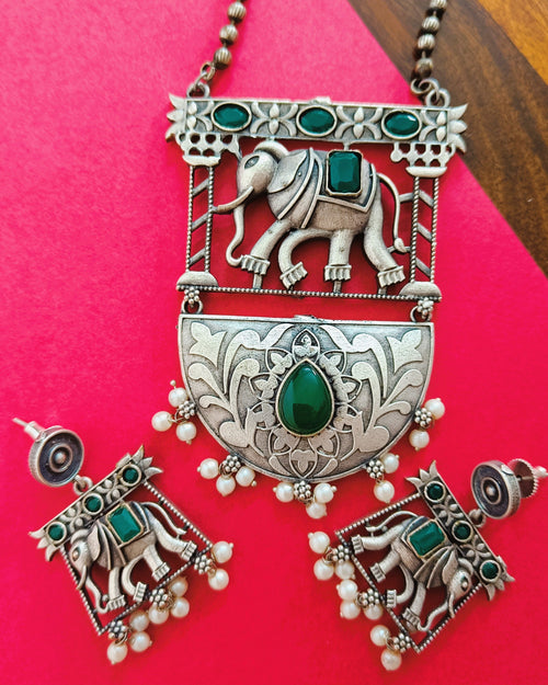 Gajraj Emerald antique oxidised silver elephant pendant necklace and stud earrings set with emerald-green stones displayed on a red background