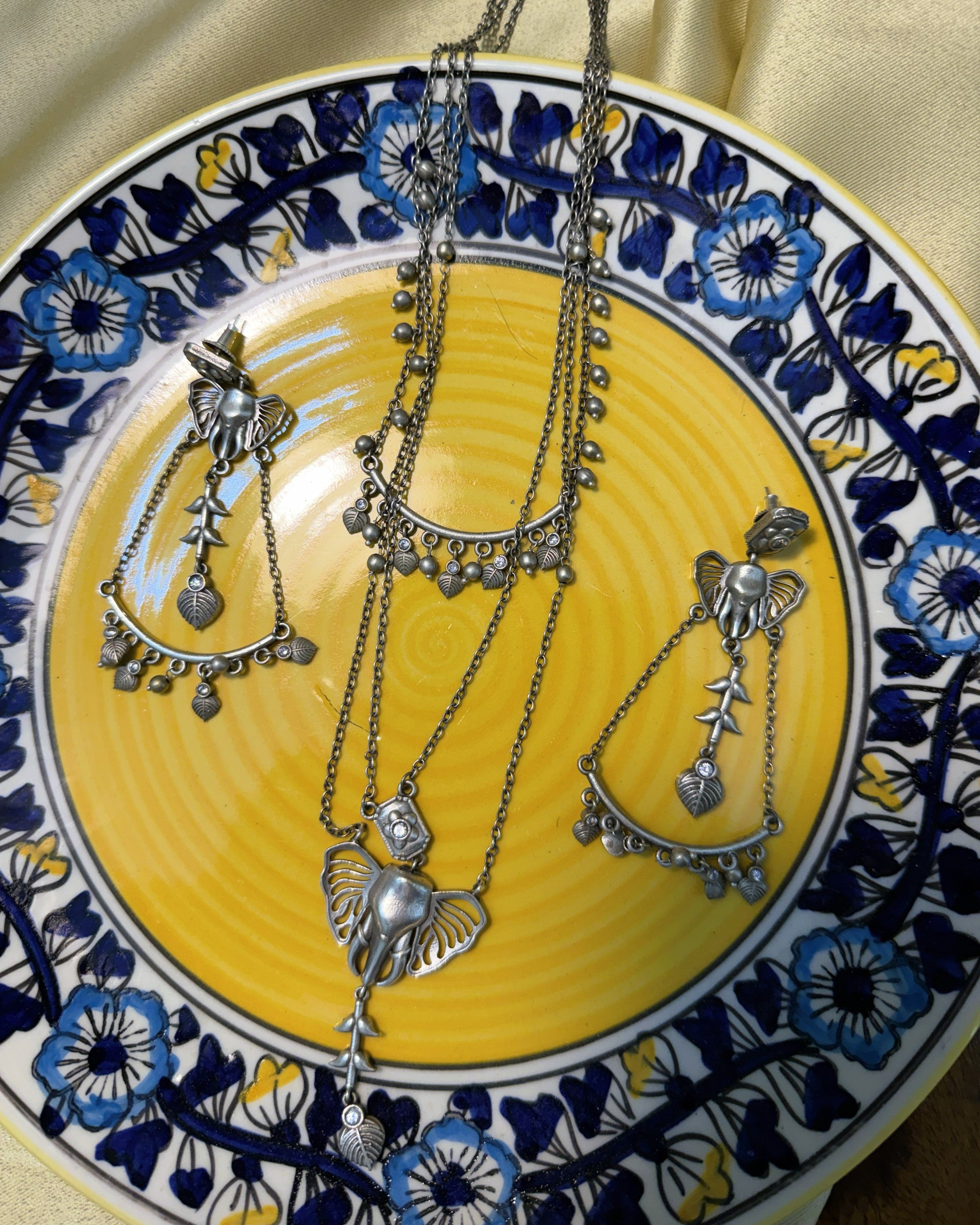 Ganesha Bloom oxidised silver necklace and earrings set displayed on a yellow ceramic plate with blue floral border