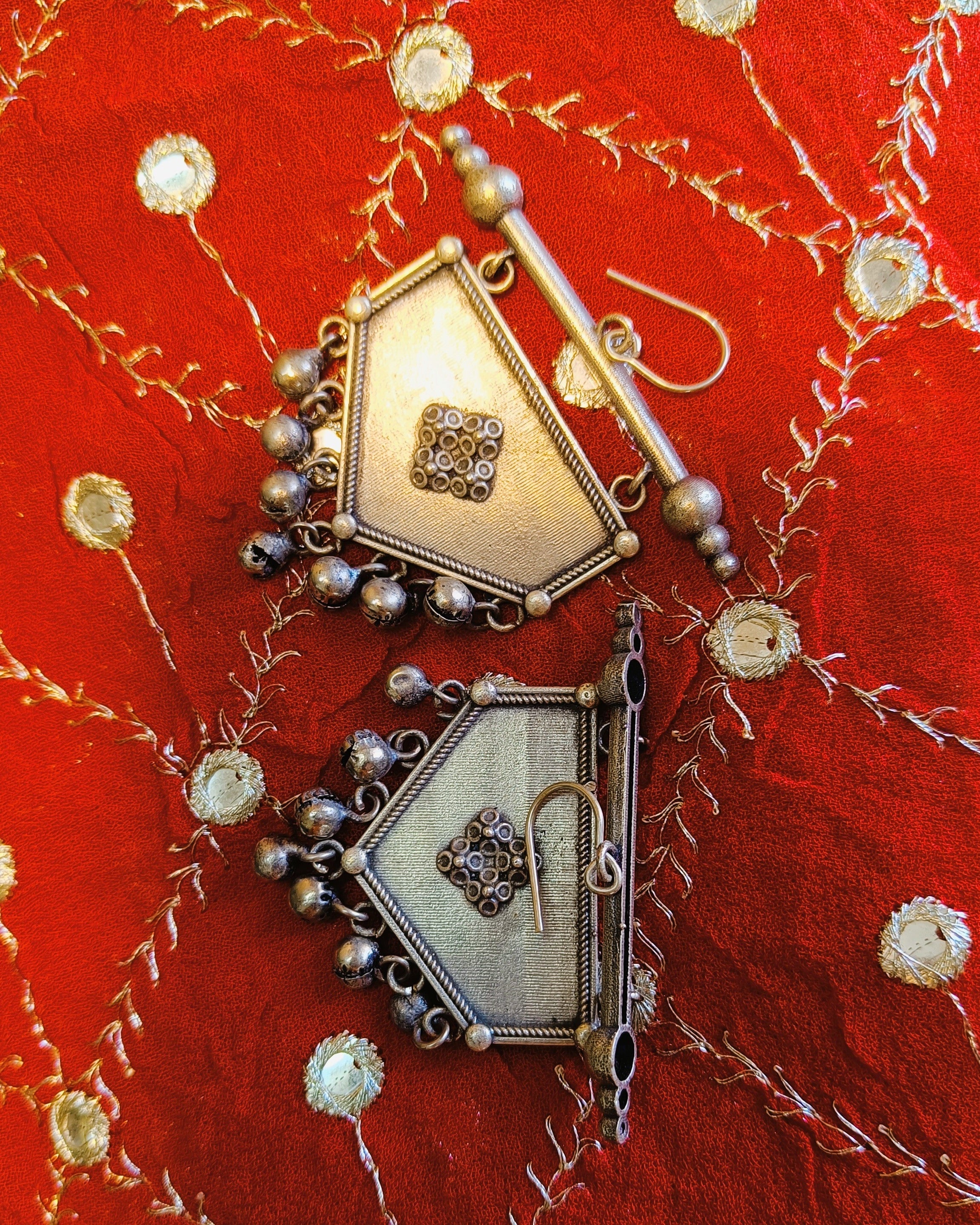 Khanjar shield-shaped antique oxidised silver drop earrings with ghungroo ball drops displayed on a red mirror-work fabric