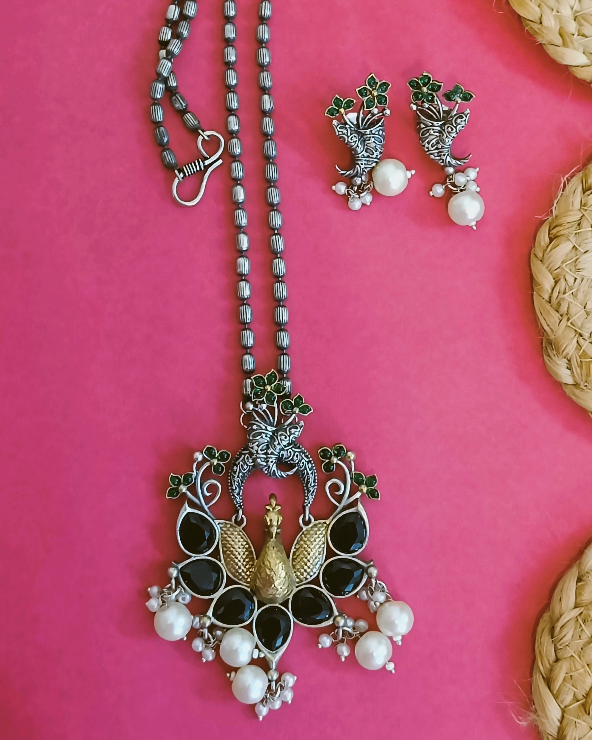 Full angled view of Mayuri Noir peacock necklace set with double-strand black bead chain, black onyx stones and white pearl drops on a red background