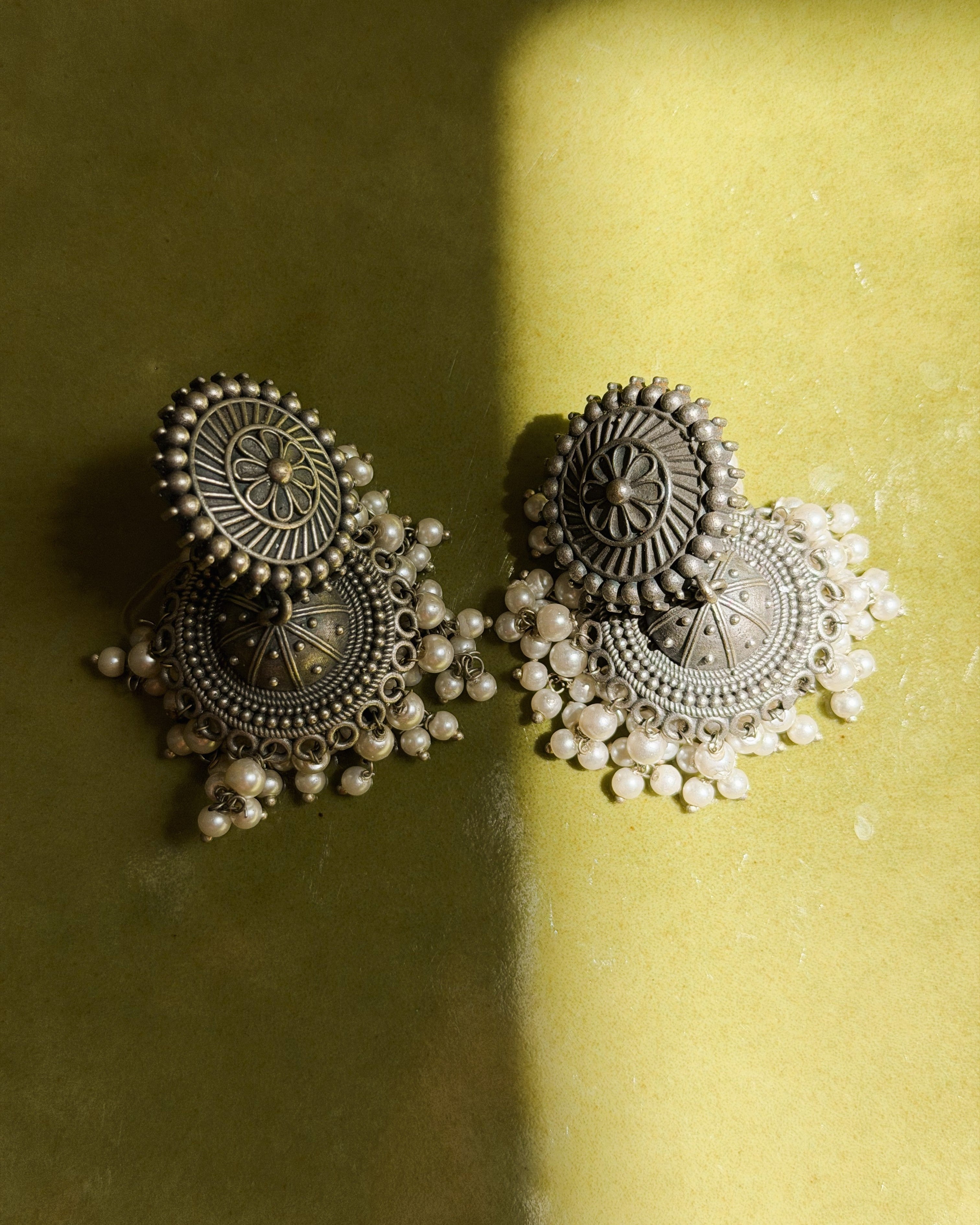 Pushpa antique oxidised silver jhumka stud earrings with floral disc top and white seed pearl drop fringe displayed on a green velvet surface