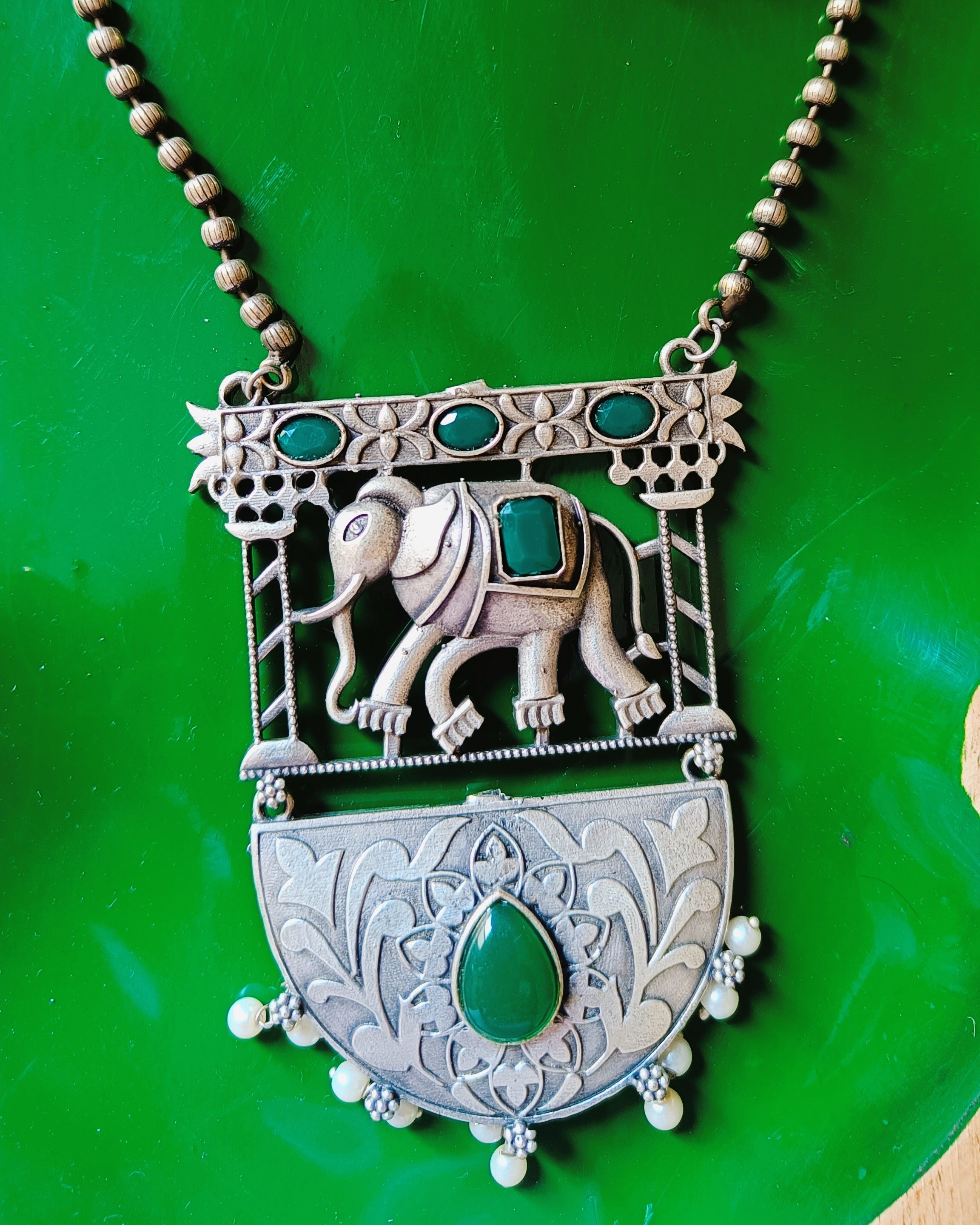 Close-up of the Gajraj Emerald two-tiered pendant featuring an elephant-in-arch motif with emerald oval stones and a lotus-engraved half-moon locket below